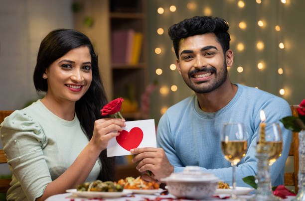Smiling Indian couple holding a red rose and a heart card at a candlelit dinner, love problem solution by astrologer Devraj Sharma